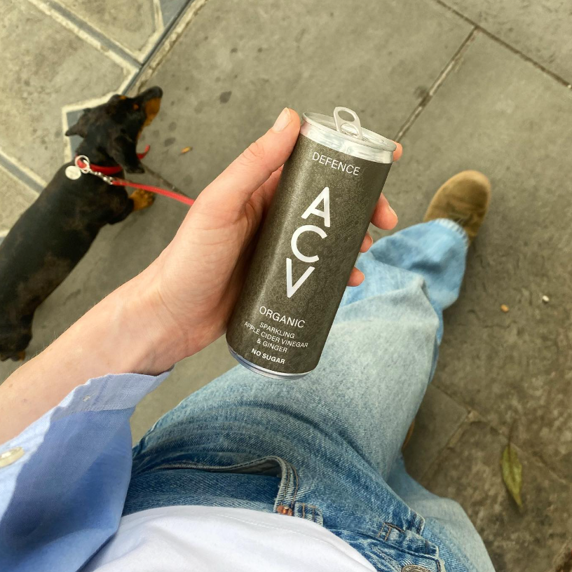 Person holding a can of Apealworld Defence ACV on a sidewalk with a dog in the background